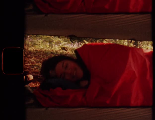A woman smiles and waves from her cozy red sleeping bag while camping in the forest on a sunny day, captured on vintage film
