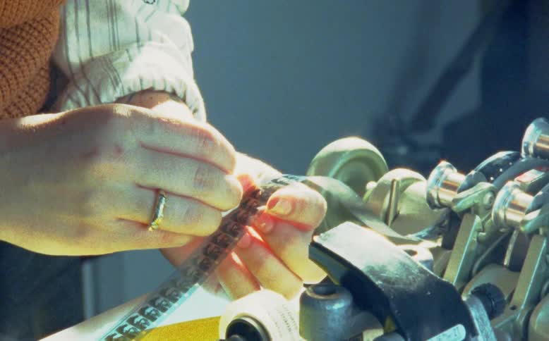 A film technician carefully cleans a strip of 8mm or 16mm movie film by hand on an editing bench