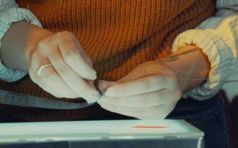 A filmmaker's hands carefully clean and inspect a strip of celluloid film on a lightbox