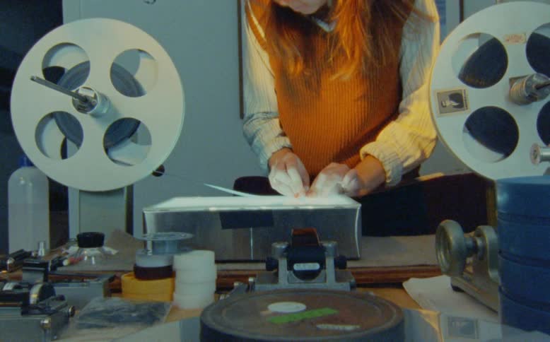 A film editor carefully inspects and cleans a strip of movie film on a vintage lightbox editing table