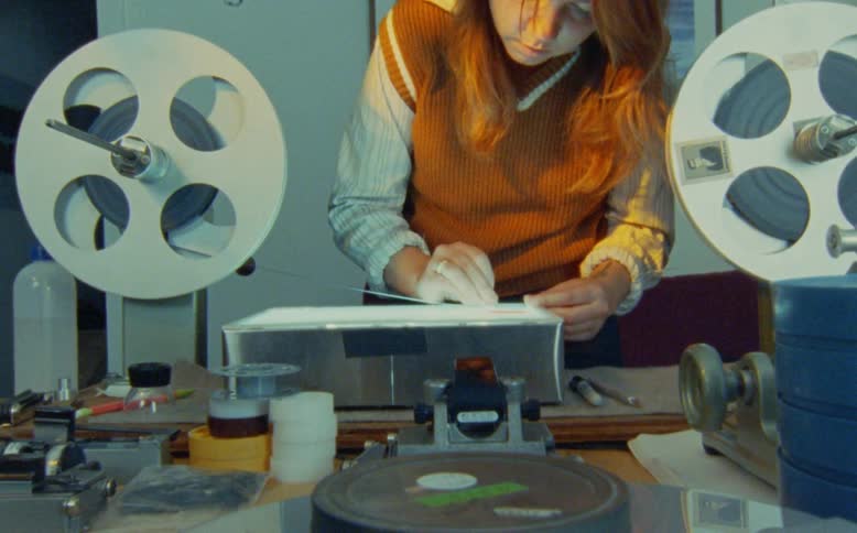 A woman in a vintage-style sweater meticulously inspects and cleans a film strip on a light box in an editing studio