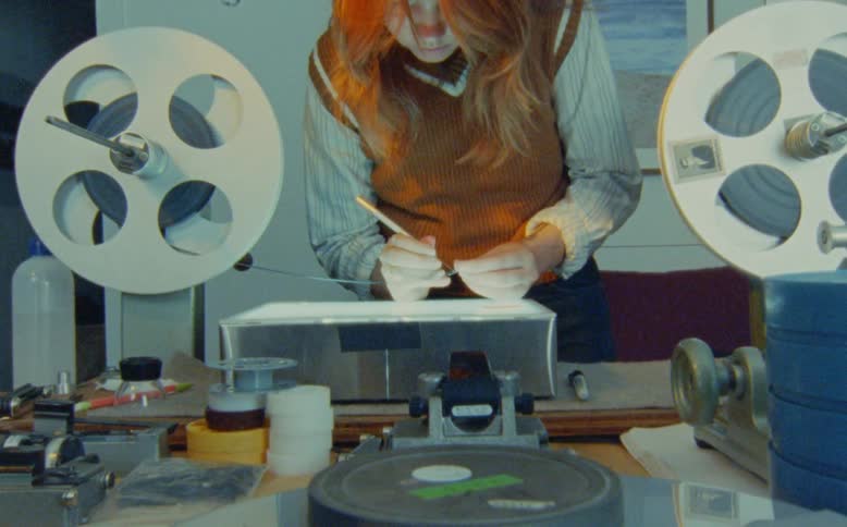 A film archivist meticulously inspects and cleans a strip of analog film on a lightbox editing bench
