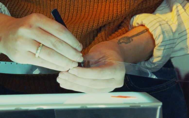 Close-up of an artist's hands carefully manipulating a strip of 16mm film with tools over a light table