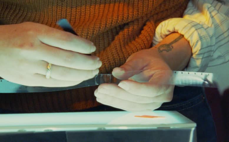 A person carefully splices a strip of vintage film together with a tool over a glowing lightbox