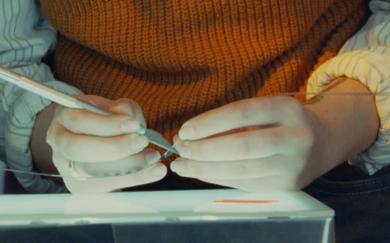 A filmmaker meticulously prepares a strip of 16mm film by hand, scraping it with a blade over a light table