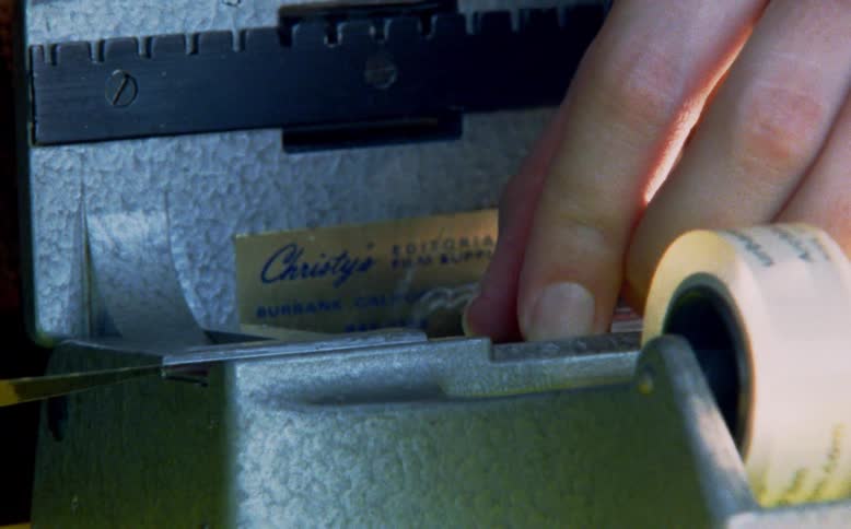 An editor meticulously splices together movie film using a vintage splicer and transparent tape