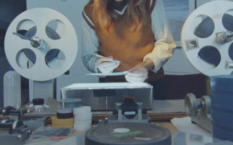 A filmmaker meticulously inspects and cleans a strip of analog film on a lightbox in a vintage editing studio
