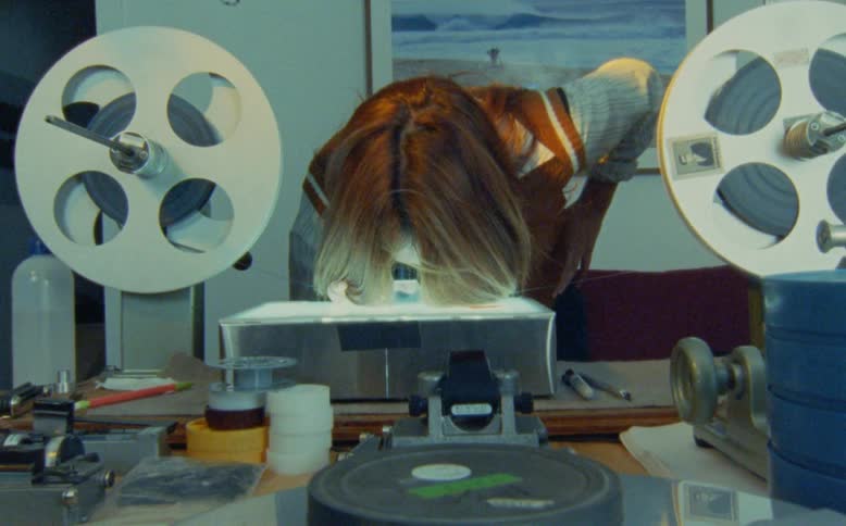 A film editor meticulously inspects a strip of 16mm or 35mm film using a loupe over a light box in a retro editing suite