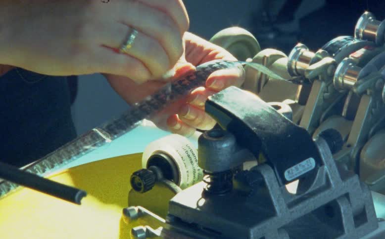 A film technician's hands meticulously clean a vintage 16mm film strip with a cotton swab next to a splicer