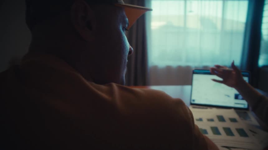 A man and woman review financial charts and data on a laptop together in a dimly lit home office