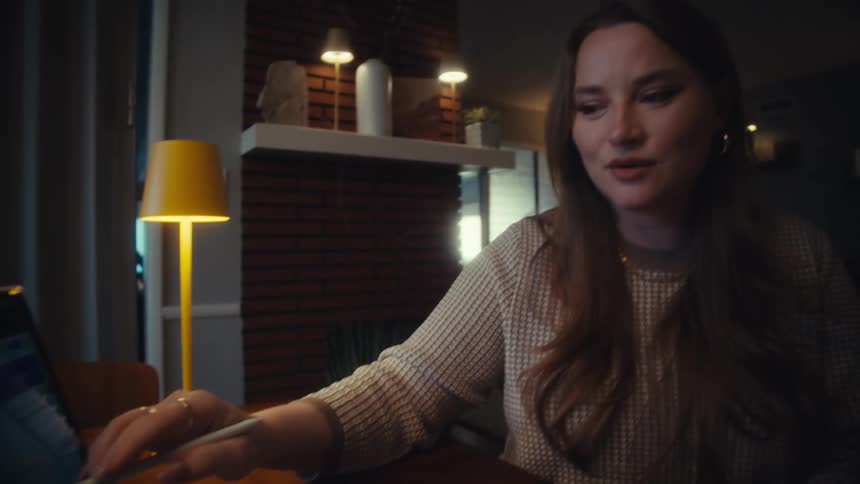 A woman working from home points a stylus at a financial chart on her laptop while discussing cryptocurrency trends at night