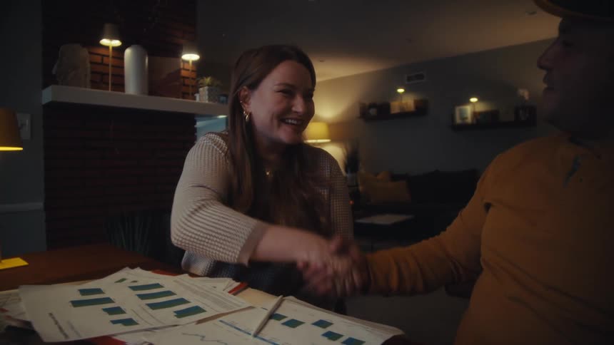 A smiling woman and man shake hands in slow motion over a table with business documents in a cozy home office