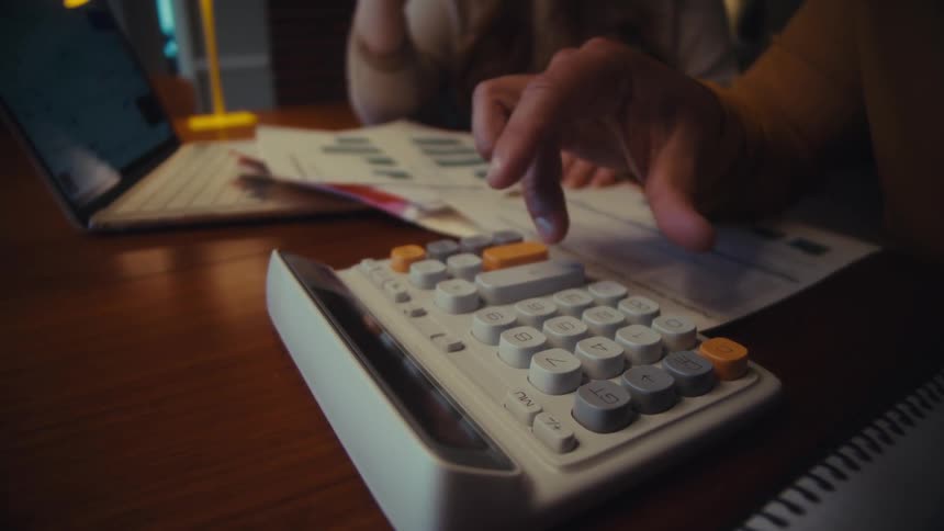 A person uses a calculator while reviewing their finances and then points to their cryptocurrency portfolio on a laptop screen