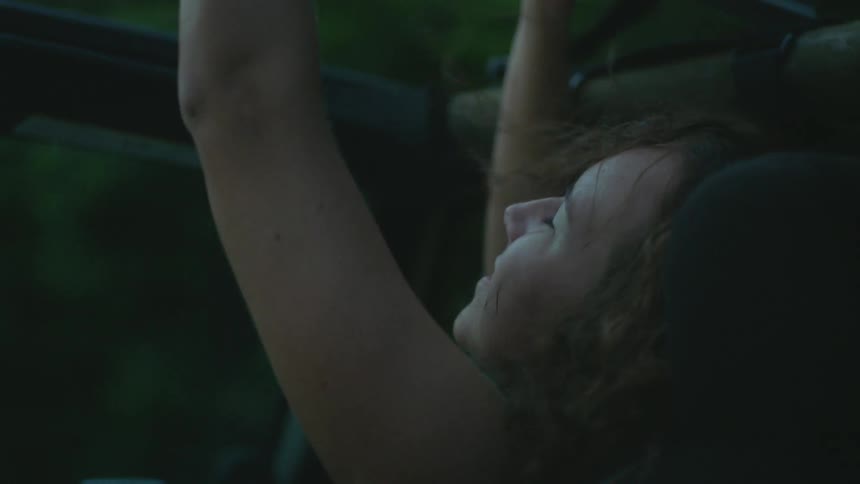 A woman rides in the passenger seat of a car with her arms reaching through the open ceiling