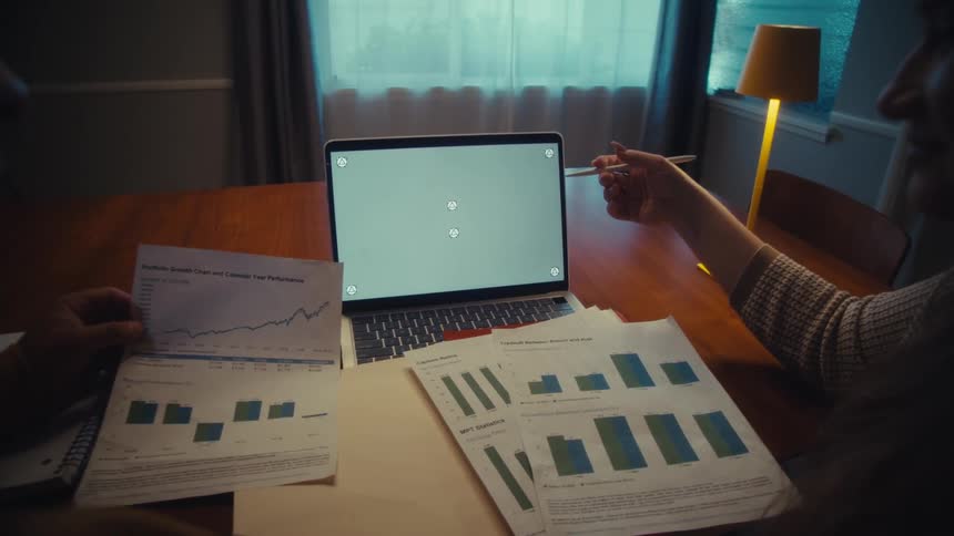 Over-the-shoulder view of two colleagues analyzing financial charts and data on papers while pointing to a laptop with a green screen