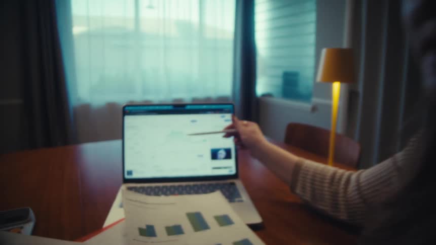 A woman points to a fluctuating cryptocurrency price chart on a laptop while discussing investments with a man at home
