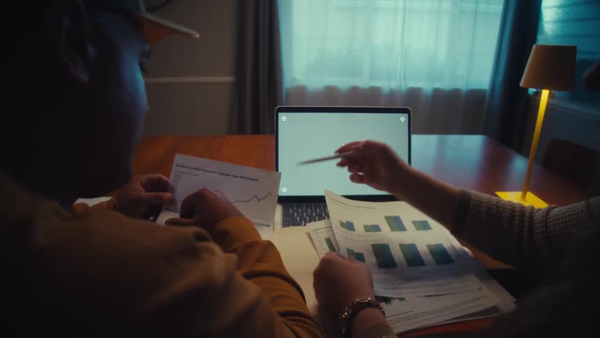 A man and woman collaborate on a project, analyzing charts on paper and a laptop with a green screen in a dimly lit room at night