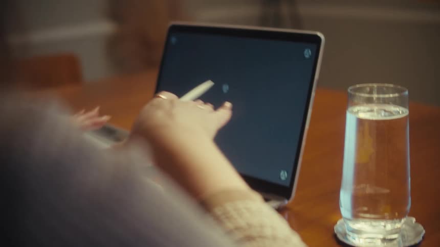 Over-the-shoulder view of a woman using a stylus and her hand to interact with a laptop's touch screen application