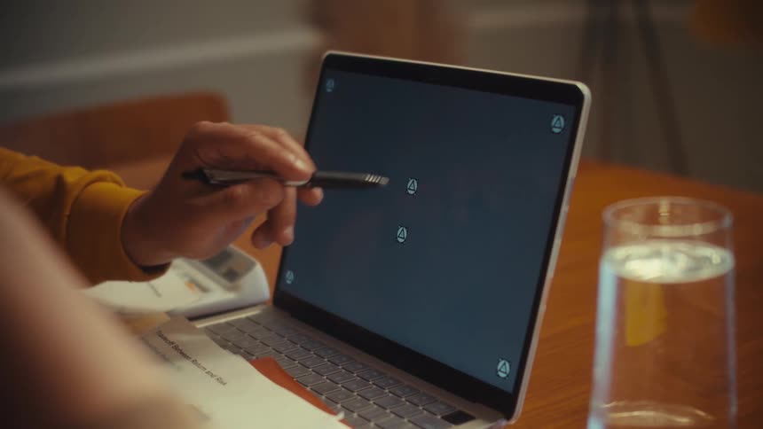 An over-the-shoulder view of a person pointing a pen at a laptop with a screen ready for compositing while collaborating with a colleague