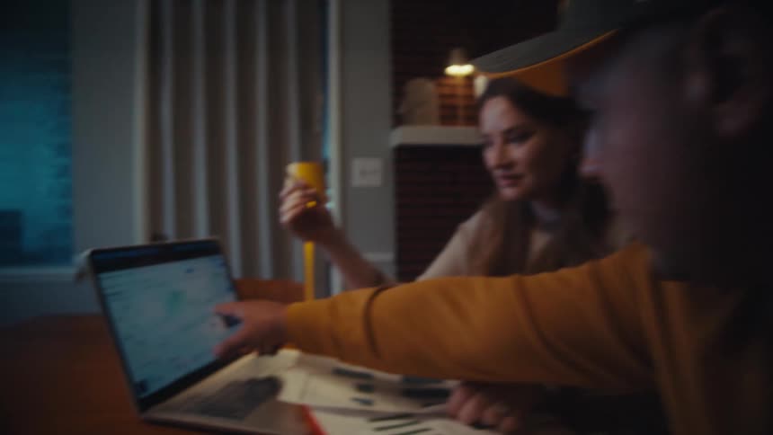 Over-the-shoulder shot of a man and woman analyzing cryptocurrency investment charts on a laptop in their home at night