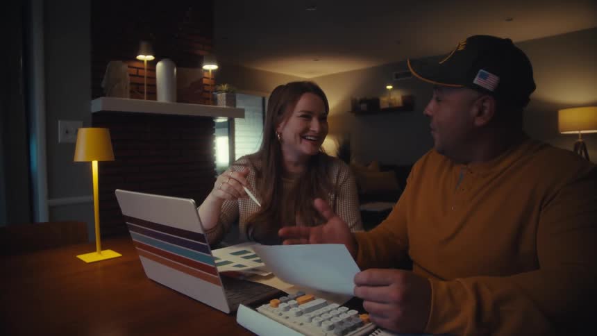 A happy veteran and his wife laugh while collaborating on their household finances at the dining table in the evening