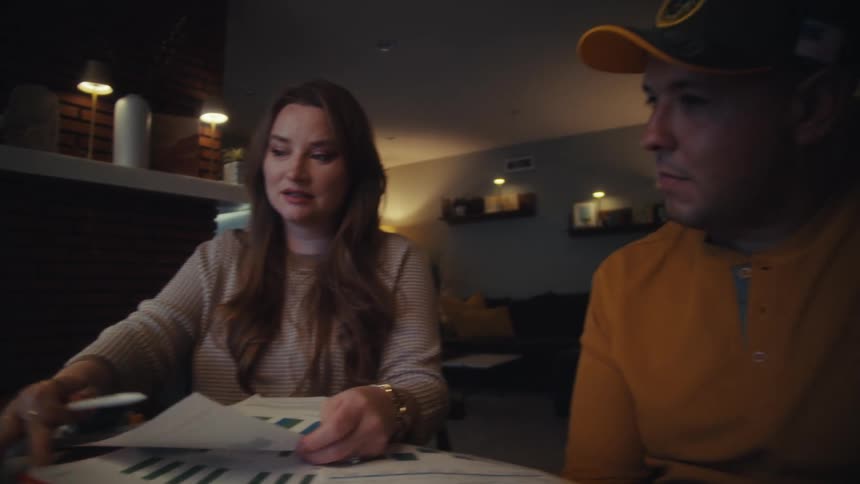 A dedicated couple works late, with the woman explaining business charts on paper and a laptop to her partner in a warmly lit home office