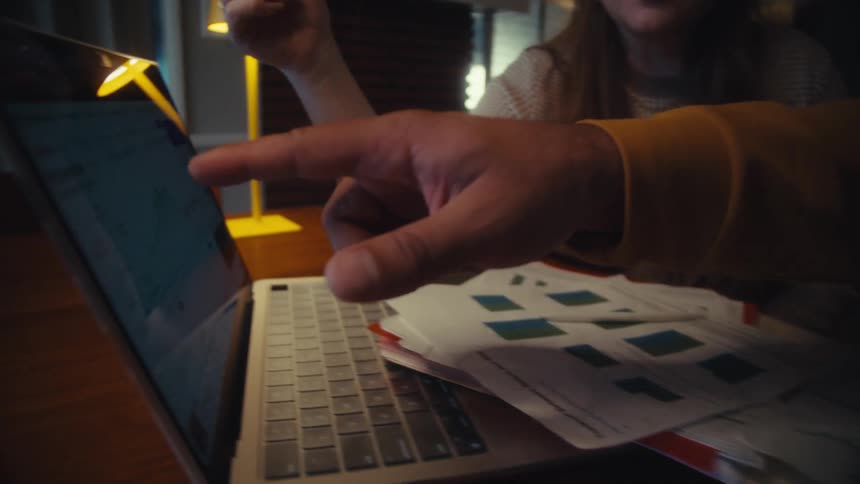 A couple reviews their investment portfolio performance by analyzing charts and graphs on paper and a laptop computer at night