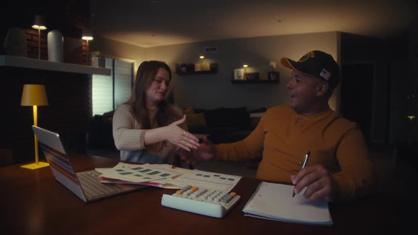 A financial advisor and her male veteran client happily shake hands, sealing a deal after a successful meeting in his home at night
