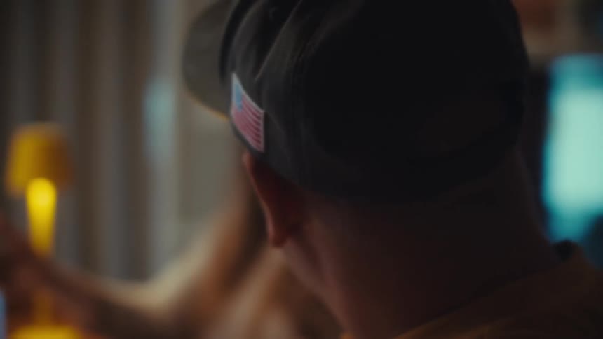A man in a baseball cap with a patriotic flag patch smiles and talks with a woman at a table in slow motion