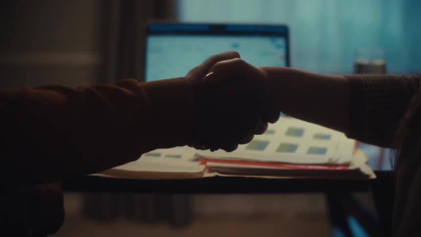 Close up of a man and woman's silhouetted hands shaking in agreement over business documents and a laptop in a dimly lit office