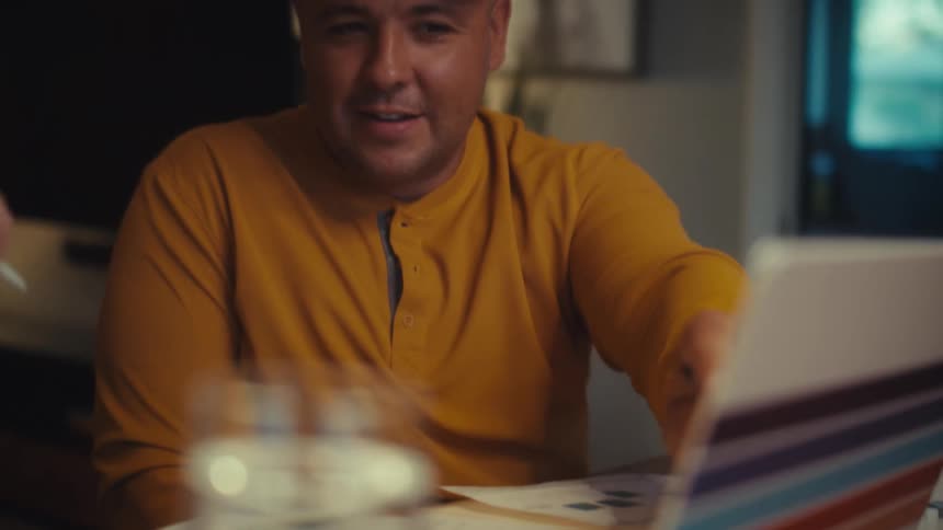 A smiling veteran in a yellow shirt collaborates with his partner at their home office, reviewing documents and using a laptop