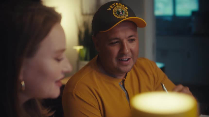 A cheerful veteran wearing a baseball cap enjoys an engaging conversation with a woman in a warmly lit room