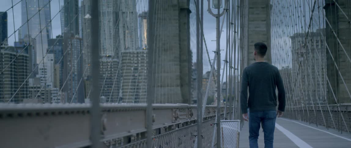 A man walks across a bridge with city skyscrapers in the distance