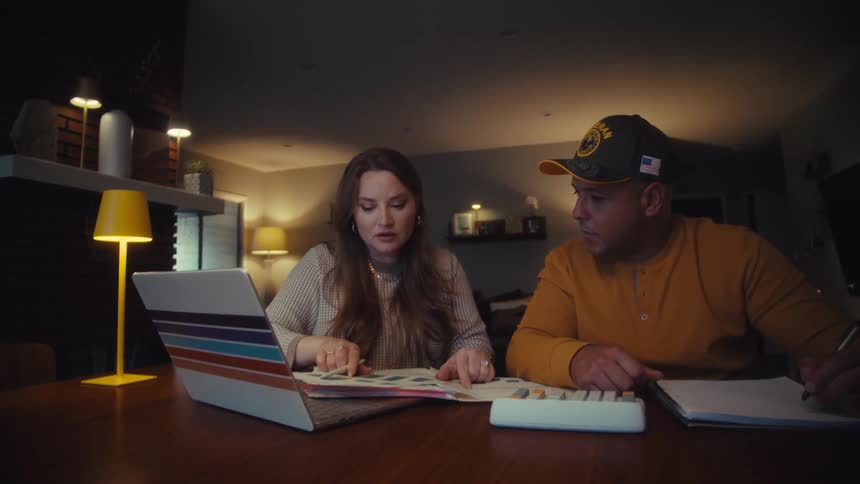 A veteran and his wife collaborate on their household budget, reviewing documents at the dining table late at night