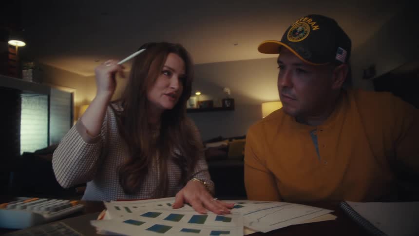 A woman explains financial charts on paper and a laptop to her veteran husband as they plan their budget together at home in the evening
