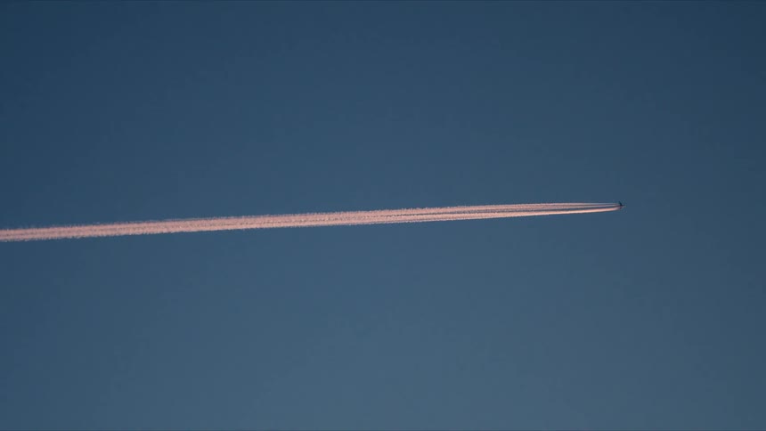 A jet airplane flies high in the atmosphere, its condensation trail glowing pink against the deep blue twilight sky