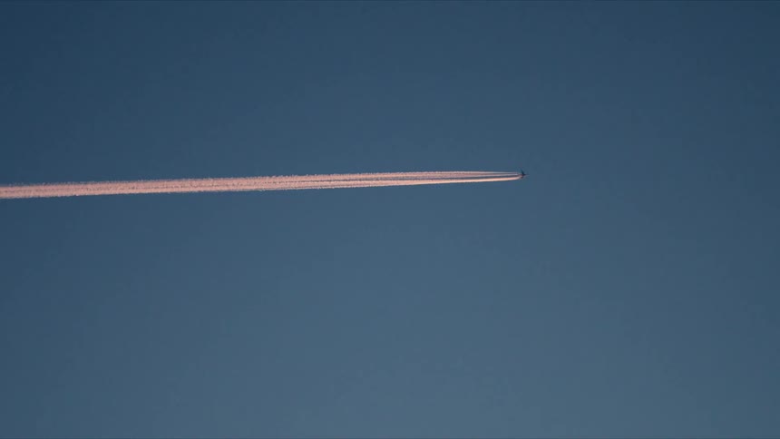 A commercial jet airplane flies high across a clear blue sky at sunset, leaving a long pink contrail in its wake
