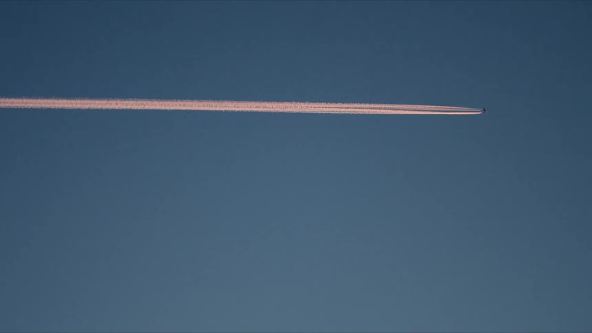 A distant passenger jet leaves a long pink contrail as it flies across a clear blue sky at sunset