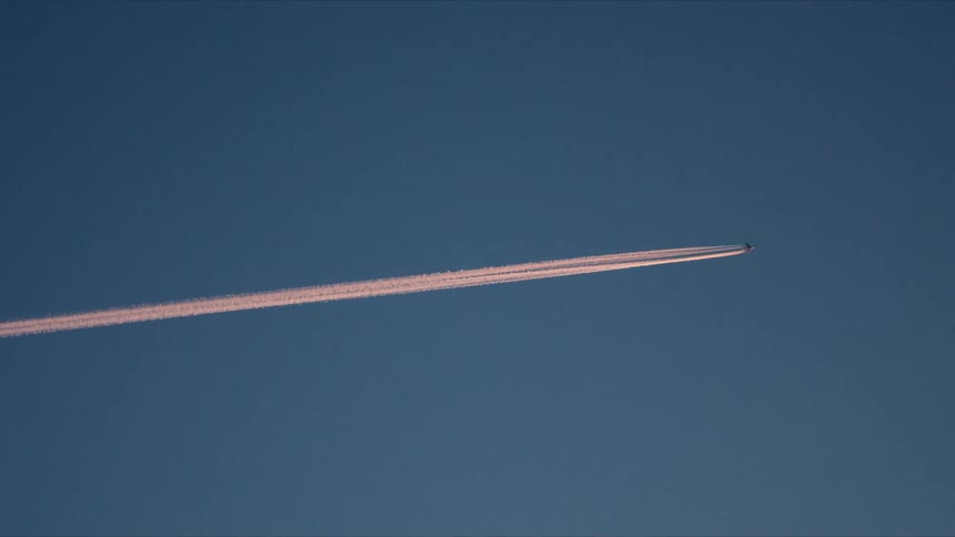 An airplane flies high in the atmosphere leaving a pink contrail against a deep blue sky during sunset