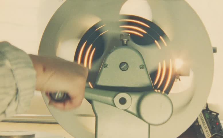 A film editor's hand manually rewinds a film reel on a vintage editing machine with a warm, hazy light flare