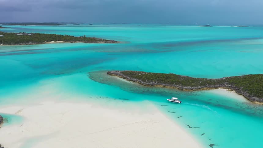 A stunning aerial view captures a boat navigating the vibrant turquoise waters and white sandbars of a tropical archipelago under a cloudy sky