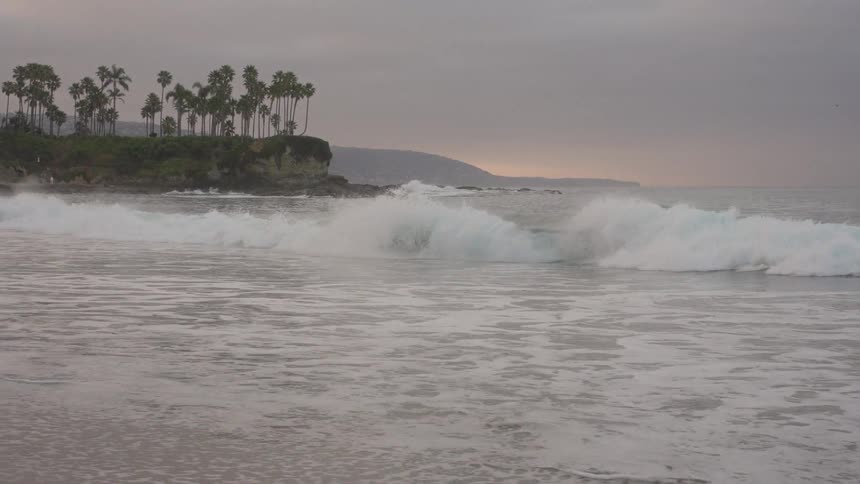 Slow-motion waves break along a picturesque coastline with palm trees silhouetted against a grey, overcast sky