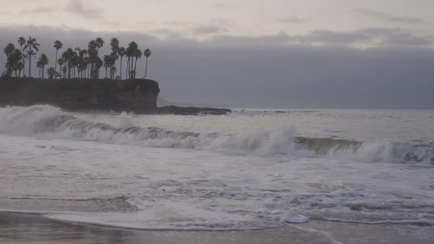 A slow-motion wave curls and breaks on a sandy shore under a cloudy sky with a cliff of palm trees in the distance