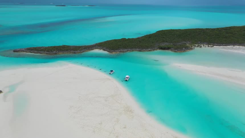 A slow aerial push-in reveals a magnificent view of a tropical sandbar, lush island, and two boats floating on vibrant turquoise ocean water