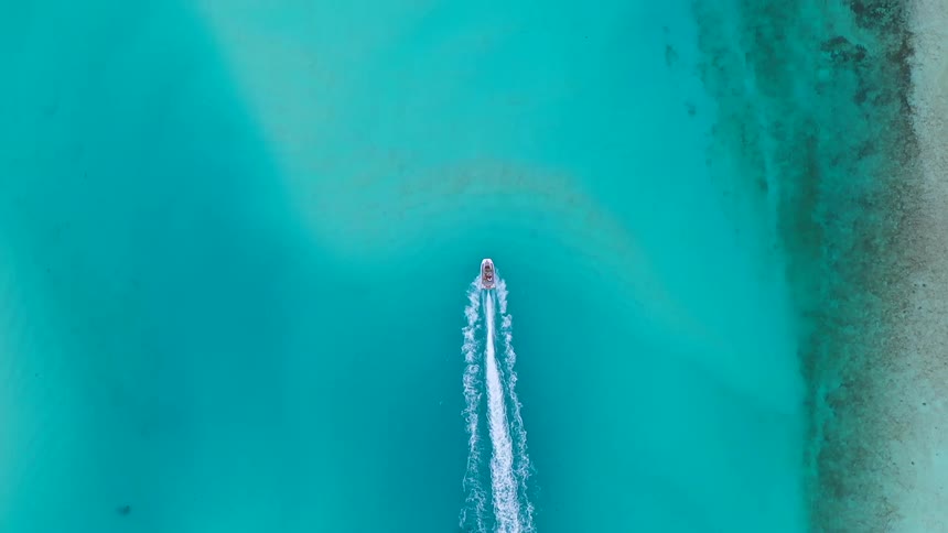 Top-down aerial drone footage shows a white boat speeding across the vibrant and clear turquoise sea