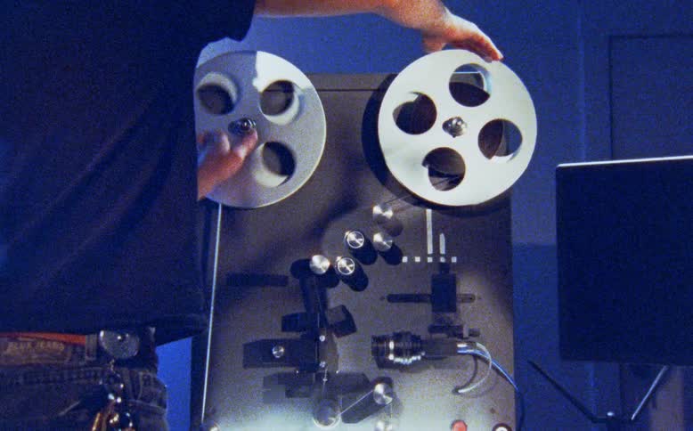 A film editor manually winds reels on a vintage flatbed film editing machine in a dark room