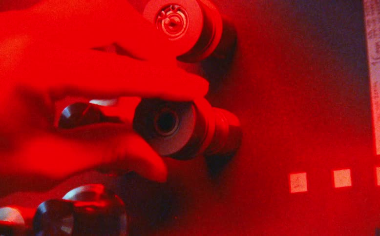 A technician operates a vintage film machine, with the scene illuminated by shifting green, blue, and red lights
