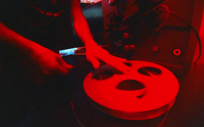 A film technician carefully loads a movie reel onto a vintage player in a dark room with shifting colored lights