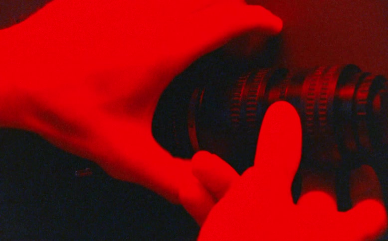 A grainy, stylized close-up shows hands adjusting a camera lens as the lighting shifts between green, blue, and red