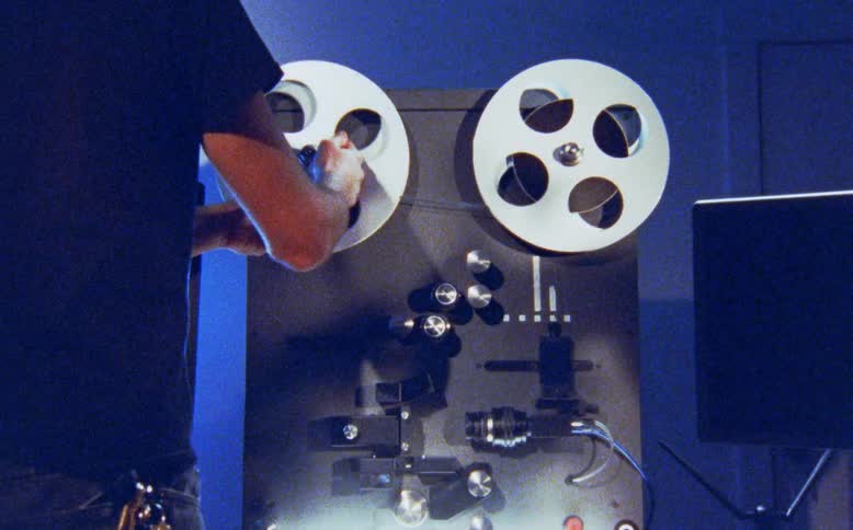 A film technician carefully threads analog film onto a reel-to-reel machine in a blue-lit editing bay